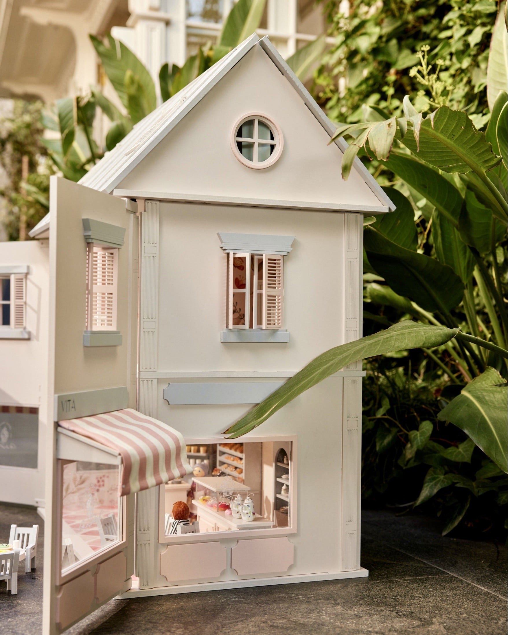 House of Rome wooden dollhouse with striped café awning, nestled among garden plants.
