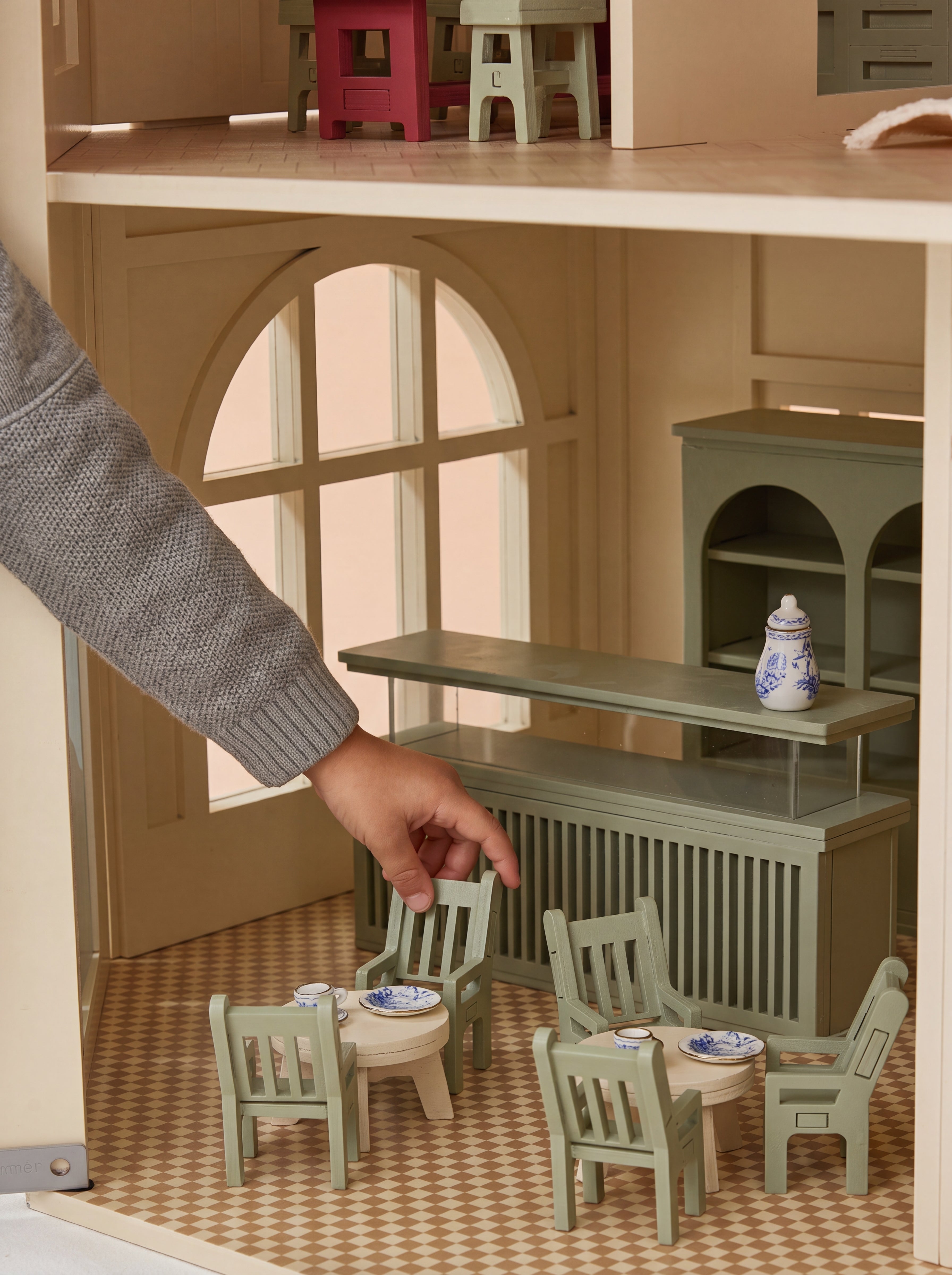 A hand arranges miniature chairs inside the House of Paris dollhouse café, highlighting the detailed interior and display counter.
