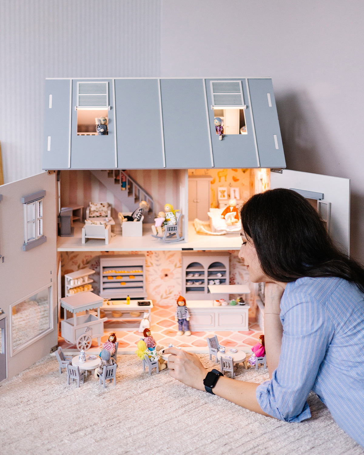 An adult arranges miniature figures inside the open House of Rome dollhouse, exploring the detailed café and bakery interior.