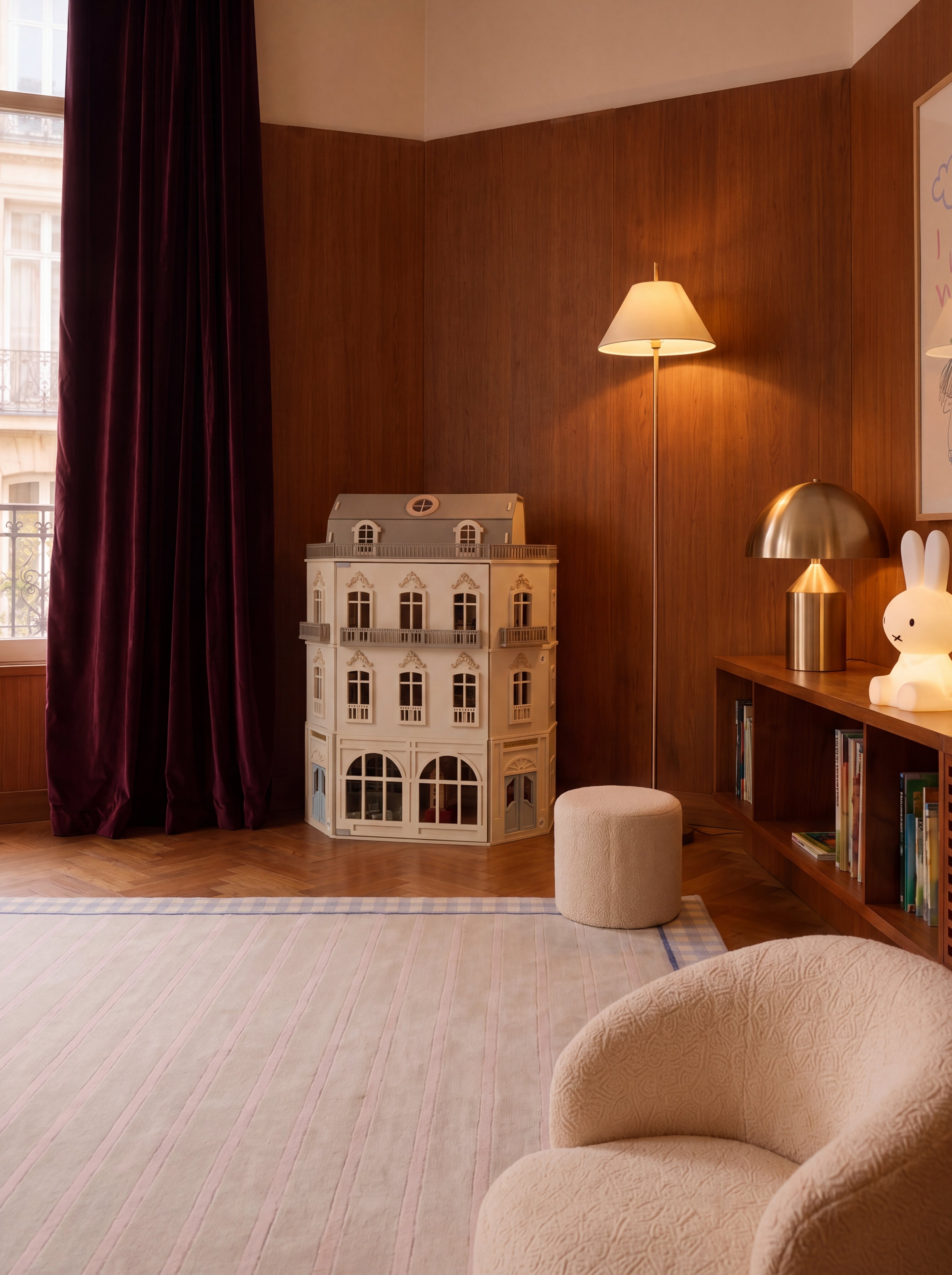 A styled interior corner with wood-paneled walls, a floor lamp, a bookshelf, a plush cream armchair in the foreground, a small cylindrical ottoman, and dollhouse and rug or play structure near a window with dark burgundy curtains.