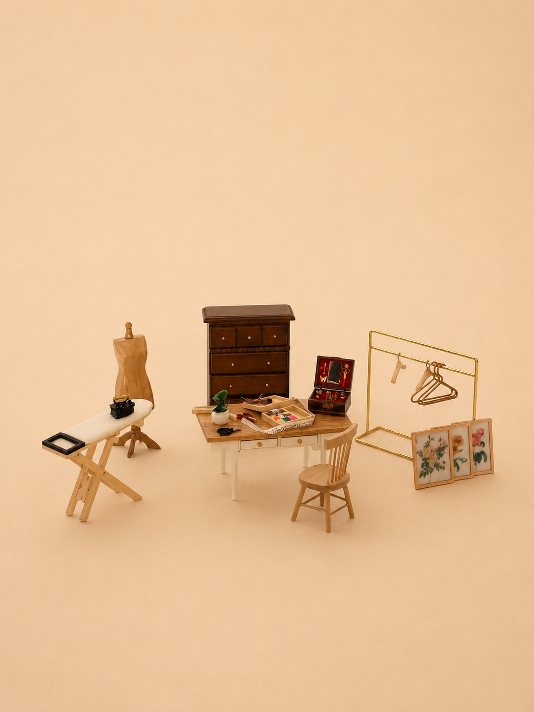 A miniature dollhouse-style room set featuring a wooden desk with a chair, a dark brown three-drawer cabinet, a gold clothing rack with hangers, a mannequin stand with a dress form, and three framed art prints, all arranged on a warm beige background.