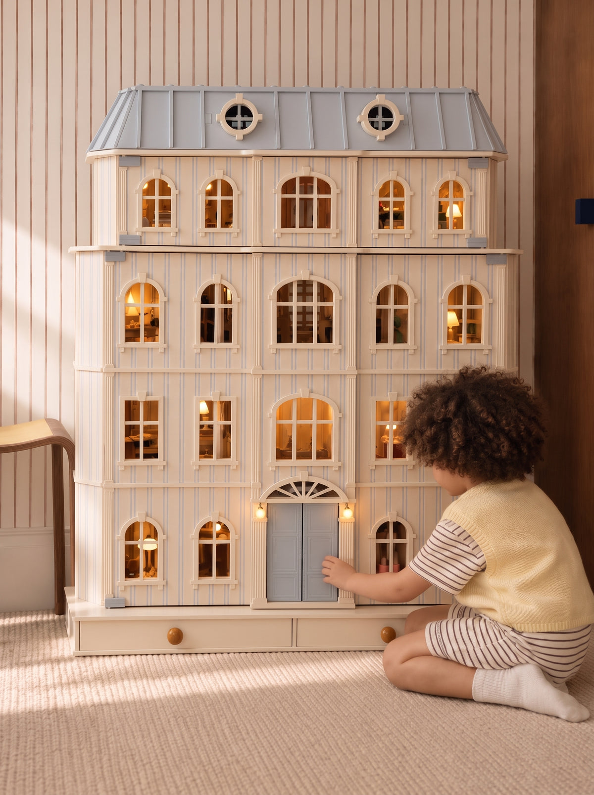 Child kneeling at the front door of the illuminated House of Monaco dollhouse against a striped wall.