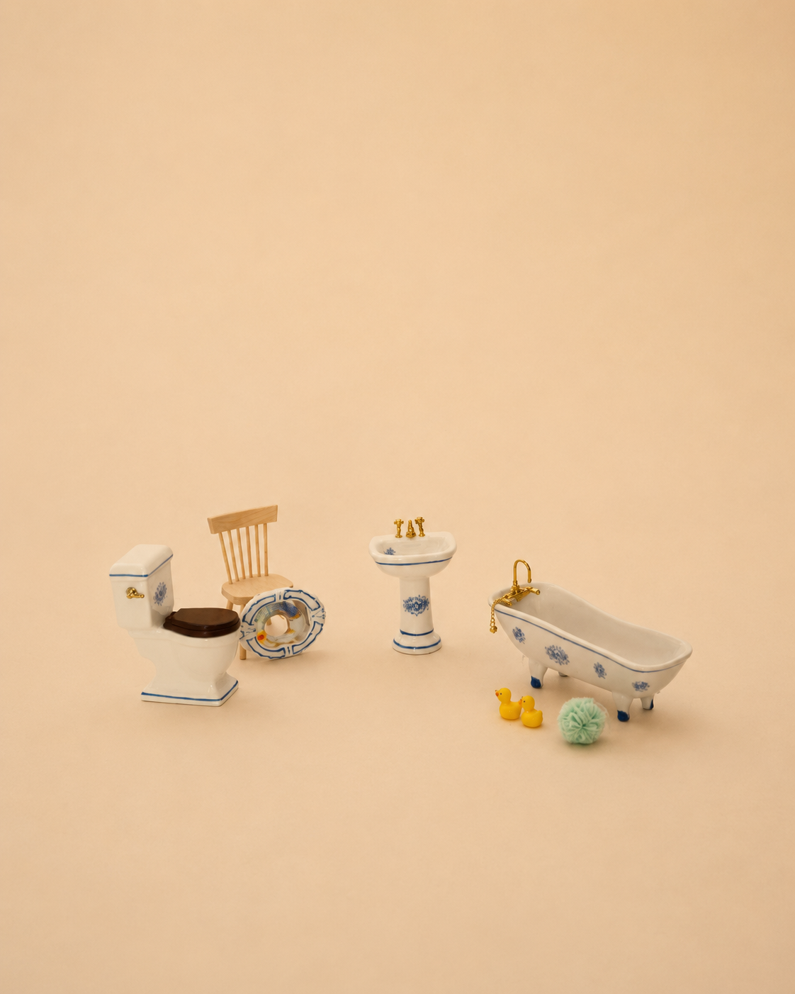 A miniature dollhouse bathroom set featuring a white porcelain toilet with a brown seat, a wooden chair, a pedestal sink with gold fixtures, a clawfoot bathtub with blue floral patterns, and small accessories, all arranged on a solid beige background.