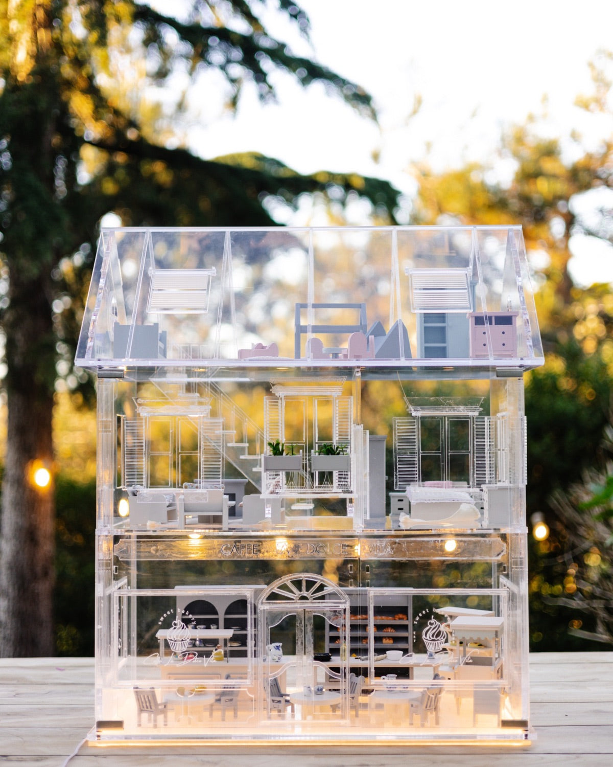 Transparent  dollhouse displayed outdoors with warm LED lighting illuminating the detailed miniature interior.