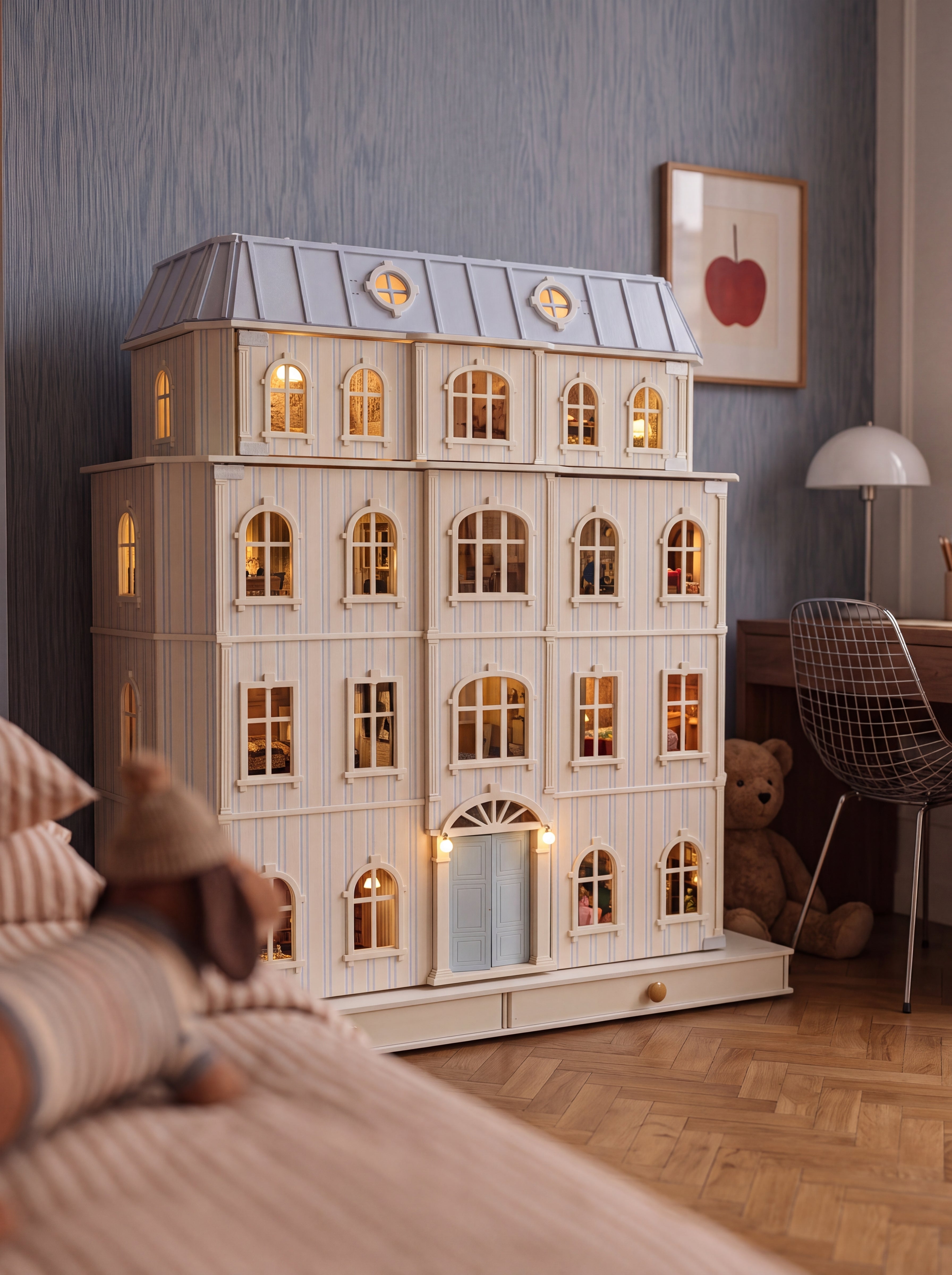 A large, multi-story dollhouse-style house with warm glowing windows, set in a styled bedroom environment with a bed, chair, and teddy bear visible.