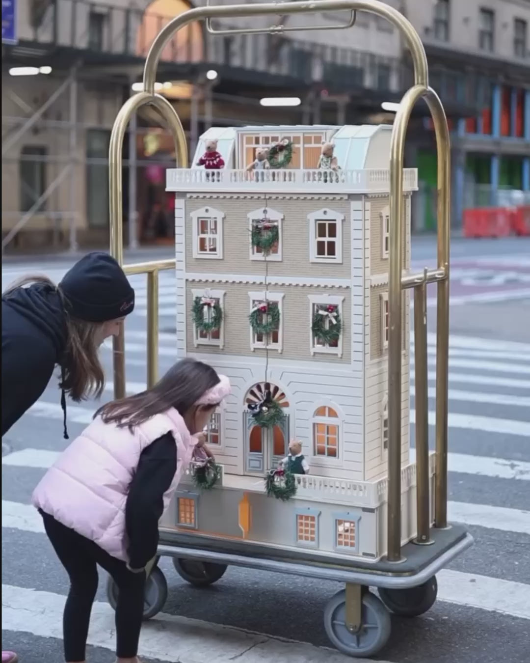 House of New York wooden dollhouse displayed on a hotel luggage cart through the streets of New York, featuring holiday decorations, Little Forest Animals doll figures, and illuminated interior windows.
