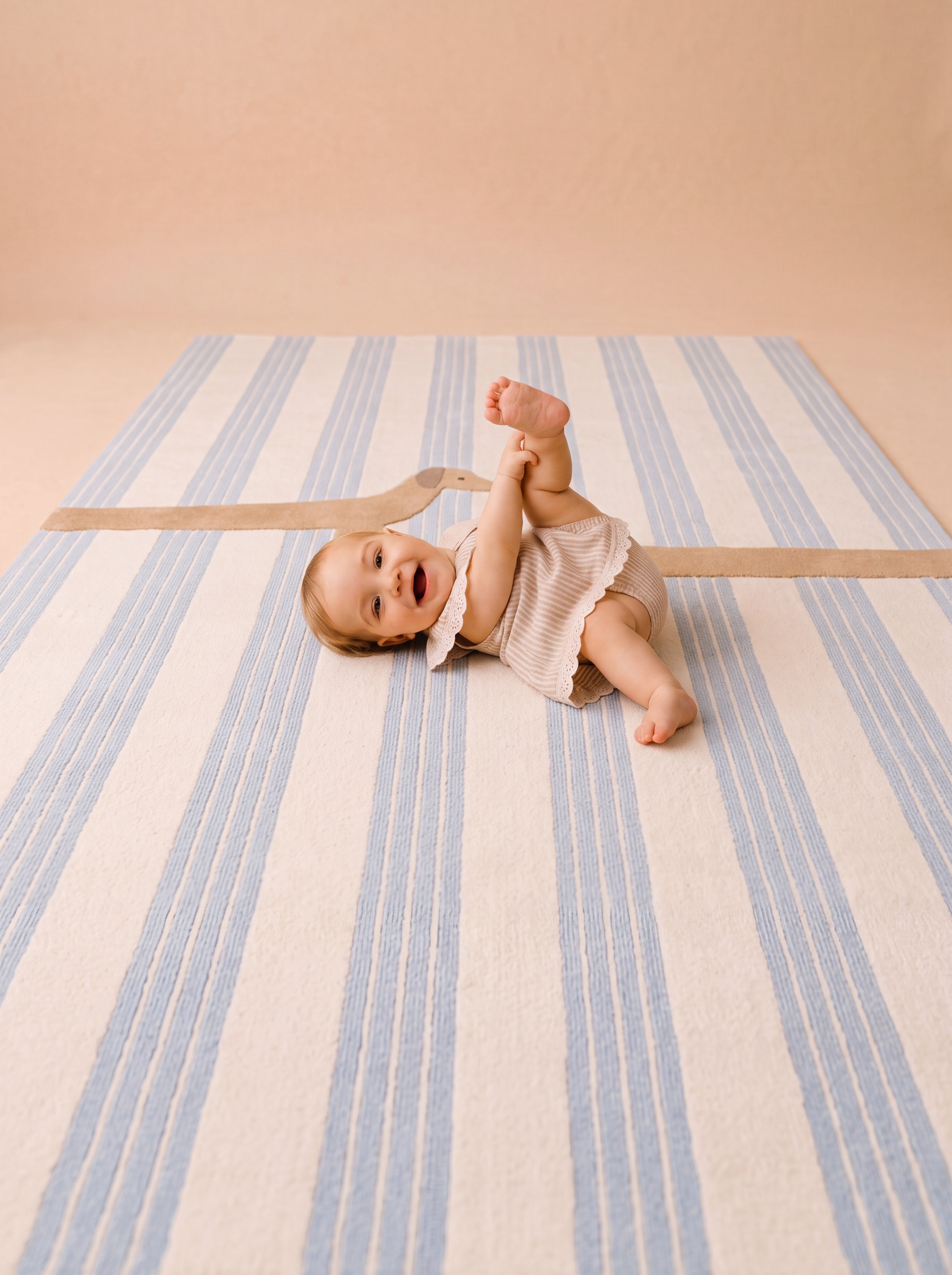 The Dachshund rug with soft blue stripes and a playful dachshund design, shown with a smiling baby lying on the rug.